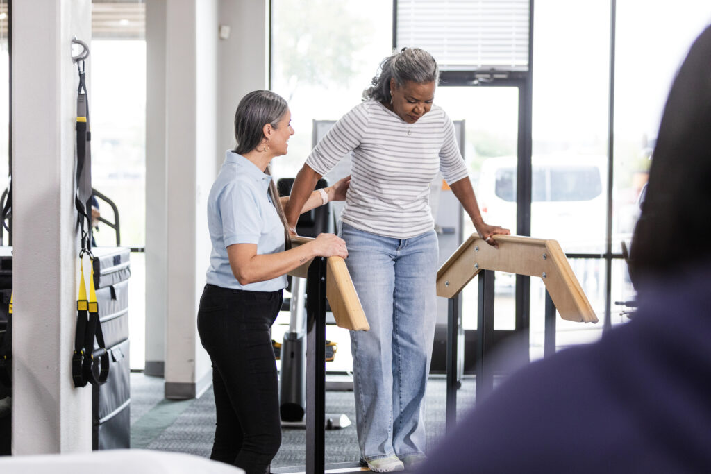 As the senior adult woman takes careful steps on the stairs at the physical therapy clinic, the mature adult female therapist reaches out to help with balance.